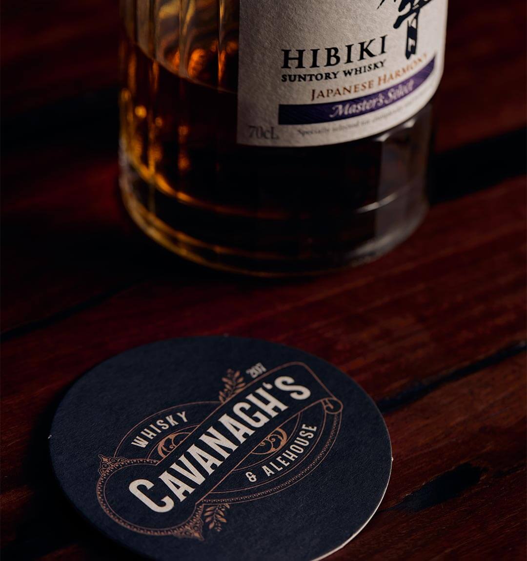 A Cavanagh's Whisky and Alehouse beer mat placed on a wooden bar next to a Hibiki bottle of Whisky.